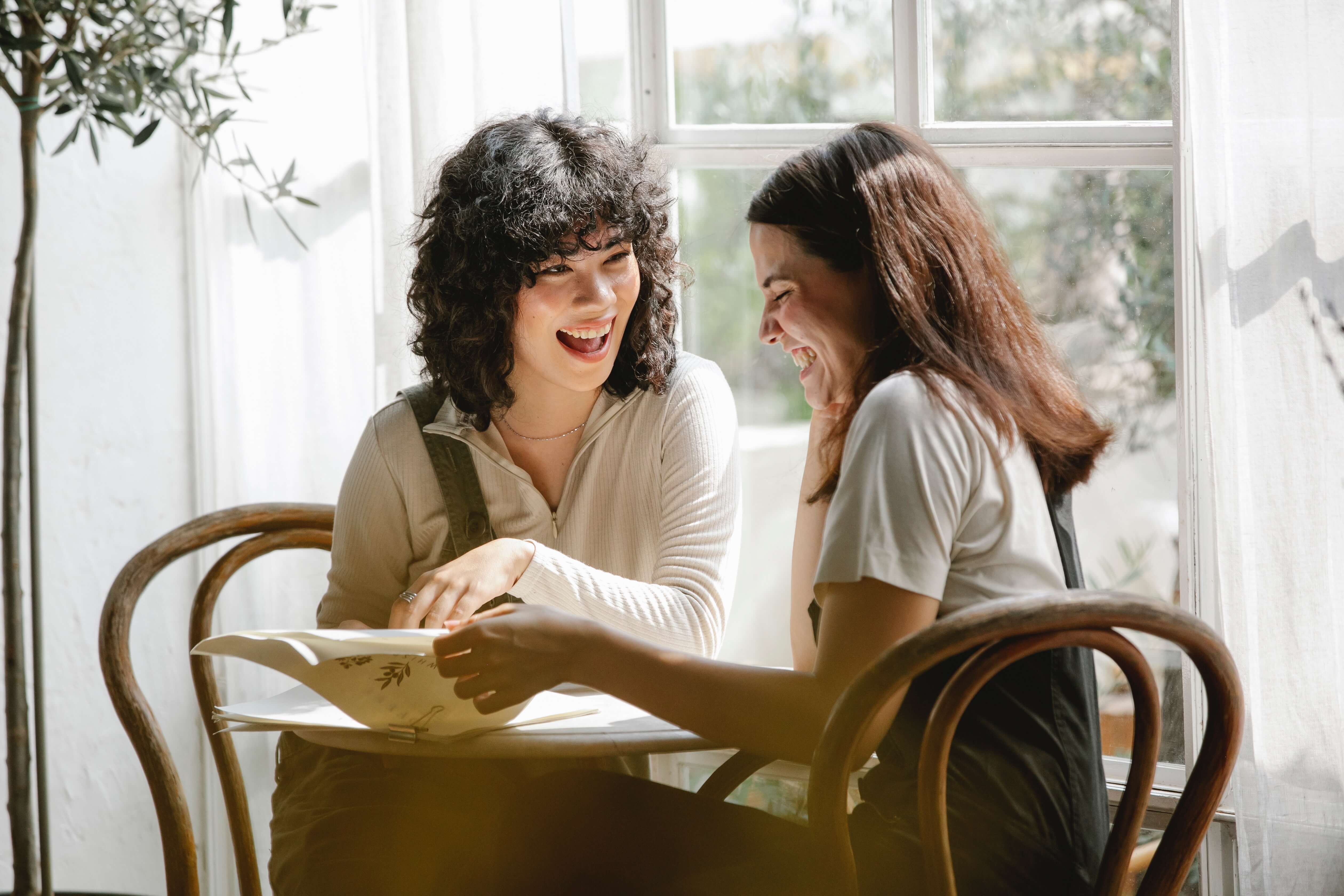 Two women sit together laughing representing the support you can find when working with a Women's Therapist in Manhattan, NY.