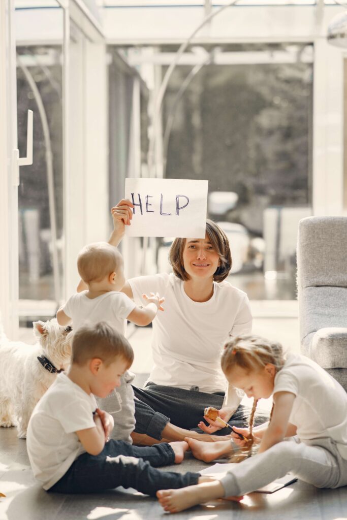 A mother sits on the floor with all three of her children feeling overwhelmed and holding a sign that says "Help" representing someone who could benefit from Therapy for Special Needs Parenting in Manhattan, NY.