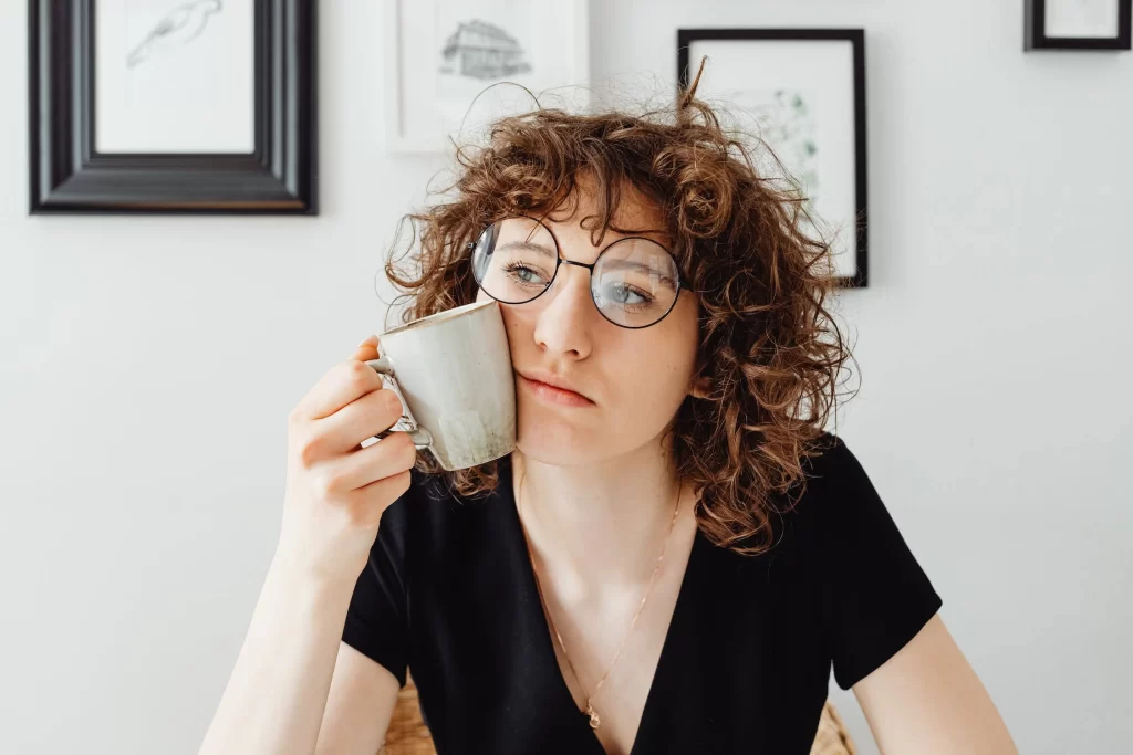 Person with curly hair and glasses thinking with a coffee cup pushed up to their face.
