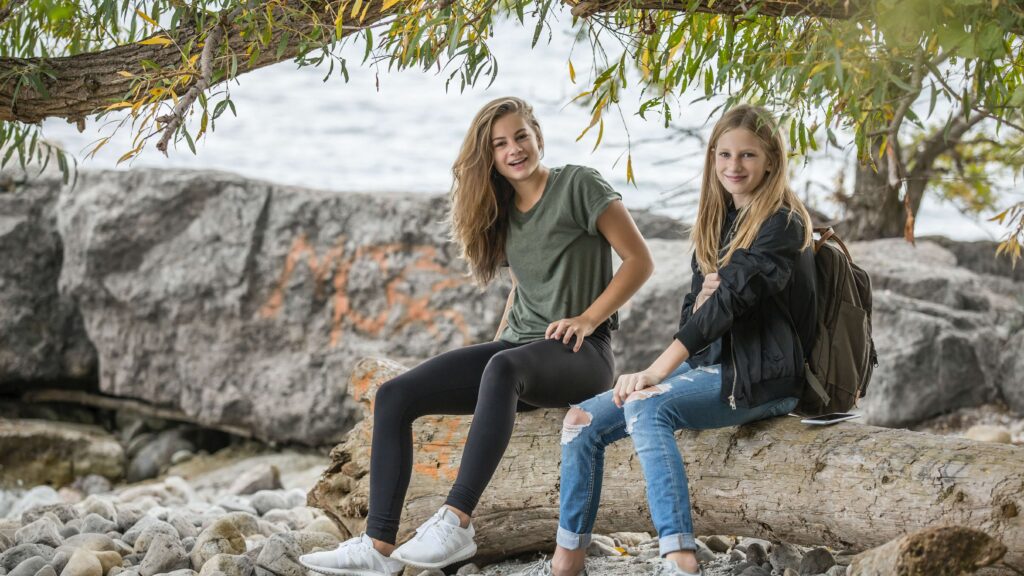 Two teenage girls sitting on rocks under a tree by water.